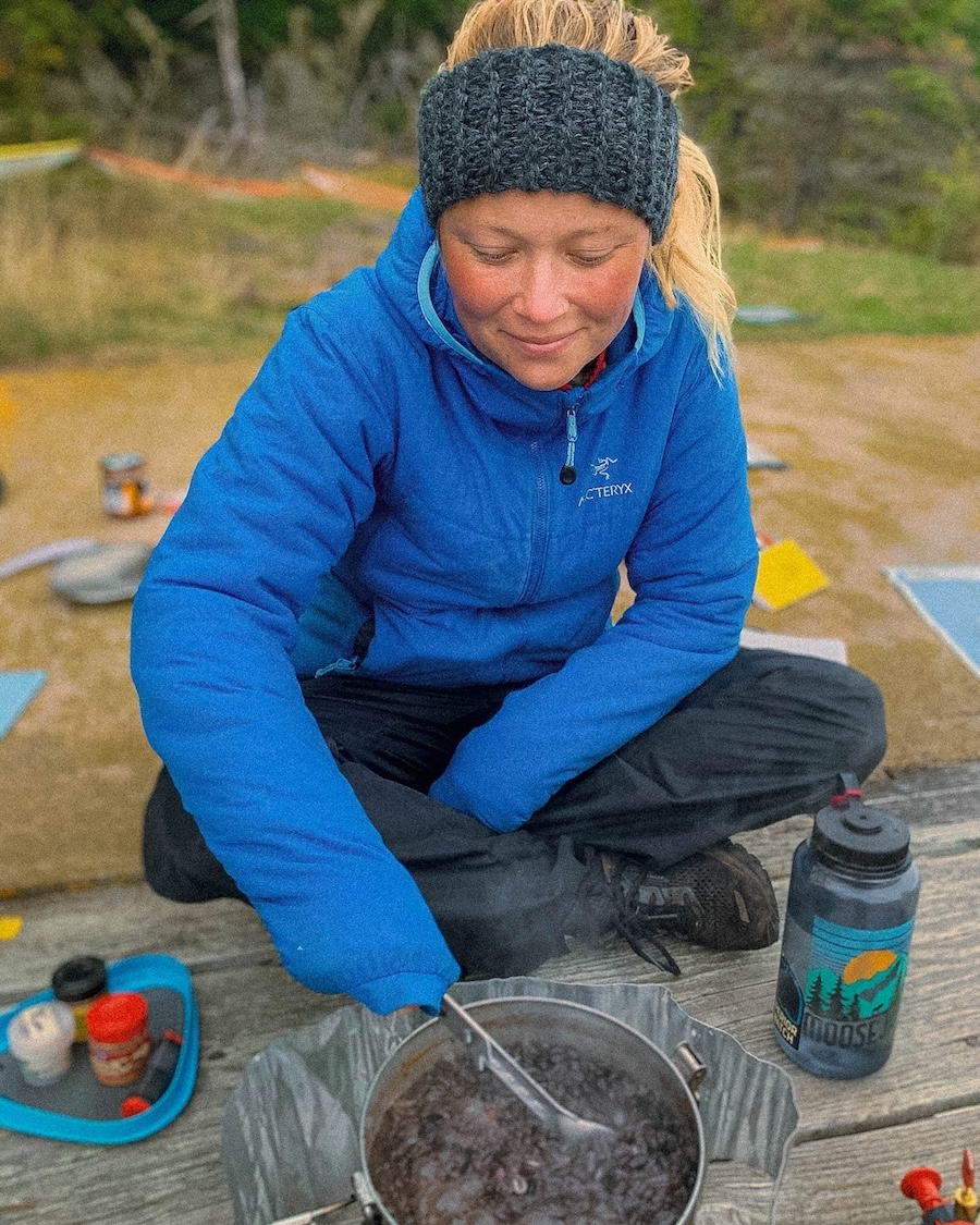 outdoor creator in a blue jacket making beans at a campfire